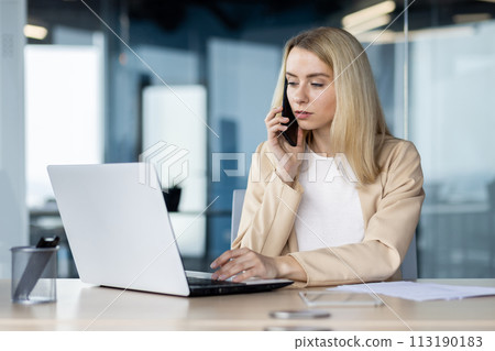 A focused blonde businesswoman in a beige blazer uses a laptop and talks on a smartphone in a modern office setting. 113190183