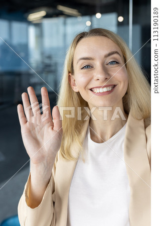 Young professional woman smiling and waving at the camera, appearing to be on a video call or taking a selfie. Young professional woman smiling and waving at the camera, appearing to be on a video call or taking a selfie. 113190189
