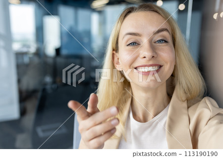 Cheerful businesswoman engaging in a video call, capturing a selfie with a smartphone camera in a modern office environment. 113190190