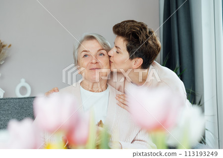 Adult woman kissing in delight, senior mom sharing a tender moment together with blurred tulips. Parenting love, care and unity concept. Mothers day. Happy family enjoying weekend together. 113193249
