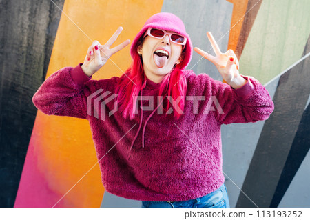 Emotional young hipster woman with pink hair and sunglasses in magenta fluffy sweatshirt and bucket hat making V sign by fingers on graffiti background. Urban street fashion. Gen Z self expression. Emotional young hipster woman with pink hair and sunglasses in magenta fluffy sweatshirt and bucket hat making V sign by fingers on graffiti background. Urban street fashion. Gen Z self expression. 113193252