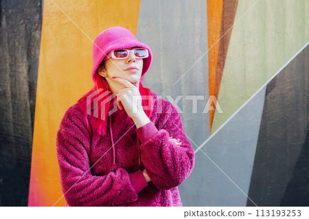 Hipster young woman with pink hair and sunglasses in magenta fluffy sweatshirt and bucket hat posing on the wall background. Urban street fashion. Mono color look. Gen Z, millennials self expression. 113193253