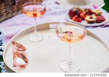 Pink drinks, cocktail with ice, raspberry, rosemary. Two glasses with martini, champagne, cider, lemonade on the blanket with fruit plate, picnic basket, Cozy summer picnic on nature. Selective focus. Pink drinks, cocktail with ice, raspberry, rosemary. Two glasses with martini, champagne, cider, lemonade on the blanket with fruit plate, picnic basket, Cozy summer picnic on nature. Selective focus. 113193259