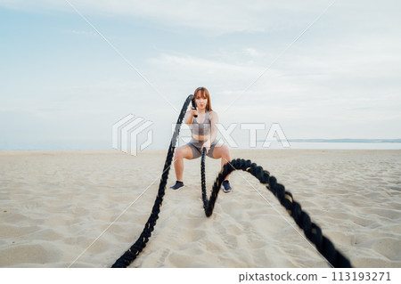 Muscular young concentrated woman working out with battle ropes on the beach, intense functional circuit training. Doing sport outdoors. Crossfit, fitness and workout concept. Selective focus 113193271