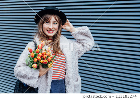 Portrait of happy, young smiling fashion woman enjoying the moment, holding bunch of fresh tulip flowers on the gray striped wall background. Urban city street fashion. Spring mood. Selective focus. 113193274