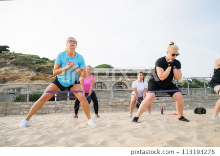 Active women of various ages doing fitness workouts in class exercise with coach on the beach. Ladies working with dumbbells and resistance bands. Sport for health and wellbeing. Active lifestyle Active women of various ages doing fitness workouts in class exercise with coach on the beach. Ladies working with dumbbells and resistance bands. Sport for health and wellbeing. Active lifestyle 113193278