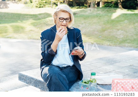 Middle-aged business woman eating sandwich burger with salad and using phone sitting on bench in the park while having lunch break. Remote work,business, freelance,blogging,social media concept Middle-aged business woman eating sandwich burger with salad and using phone sitting on bench in the park while having lunch break. Remote work,business, freelance,blogging,social media concept 113193283