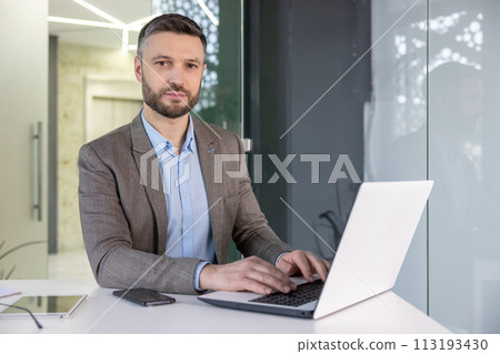A focused professional man in a suit working intently on a laptop in a modern office setting, exuding a serious demeanor. 113193430