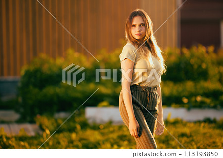 26 year old woman stands in front of wall of wooden house at sunset, mid shot portrait outdoor portrait. 26 year old woman stands in front of wall of wooden house at sunset, mid shot portrait outdoor portrait. 113193850