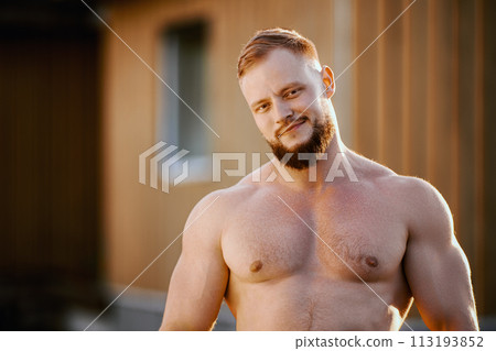 Ironic athlete with goatee without shirt outdoors near wooden rural house outside city. Ironic athlete with goatee without shirt outdoors near wooden rural house outside city. 113193852