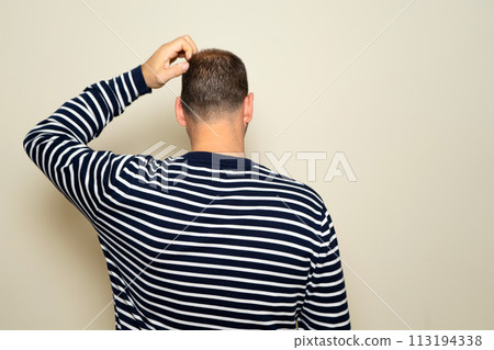 Young Hispanic man scratching his head with his back to the camera, he has doubts about how to solve a problem. Isolated on beige studio background. 113194338