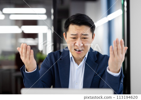 An Asian businessman looking frustrated and confused at his computer in a modern office setting. 113194620