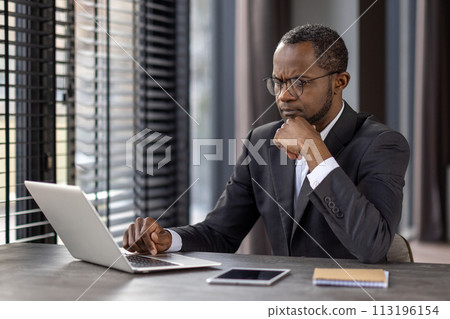 A professional businessman in a suit concentrating on his work on a laptop at a modern office desk, possibly analyzing data or planning strategies. 113196154