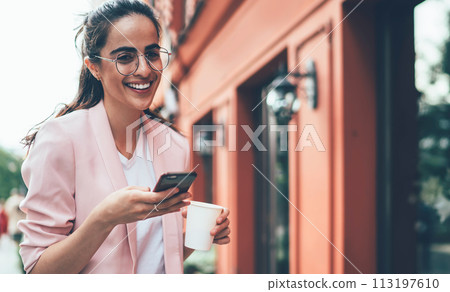 Cheerful student with takeaway coffee using smartphone on street in downtown 113197610