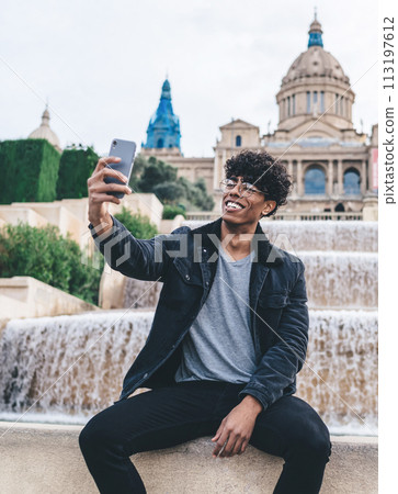 Cheerful black man taking selfie at fountain edge with cellphone 113197612
