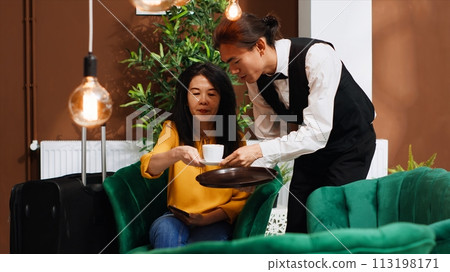 Male waiter bringing coffee to woman in lounge area, waiting to go through with check in process. Bellhop serving drink for hotel guest, providing great services at resort. Handheld shot. 113198171