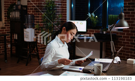 Asian employee reluctantly doing work in office late at night, bored of checking financial data charts on laptop. Businesswoman annoyed by having to check paperwork, feeling stressed and overworked 113198183