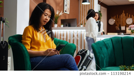 Traveler searching landmarks and places to visit during her stay, using smartphone and relaxing in lounge area at hotel. Asian woman creating a plan for the vacation, waiting to confirm booking. Traveler searching landmarks and places to visit during her stay, using smartphone and relaxing in lounge area at hotel. Asian woman creating a plan for the vacation, waiting to confirm booking. 113198214