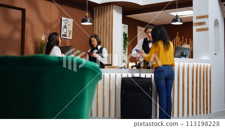 Front desk staff assisting women with registration process, asking to see passport and sign confirmation documents for accommodation. Two guests arriving at five star holiday retreat. 113198228