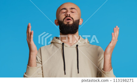Muslim person presenting his belief in Allah by praying on camera, looking up and saying a prayer for forgiveness. Middle eastern guy worshiping God with faith, religious man in studio. Camera 2. 113198271