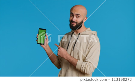 Muslim guy holding smartphone with greenscreen display, pointing at isolated chromakey template on mobile phone app. Young adult presenting blank copyspace mockup layout. Camera 1. Muslim guy holding smartphone with greenscreen display, pointing at isolated chromakey template on mobile phone app. Young adult presenting blank copyspace mockup layout. Camera 1. 113198273