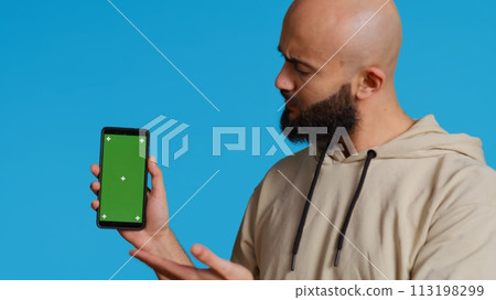 Muslim guy holding smartphone with greenscreen display, pointing at isolated chromakey template on mobile phone app. Young adult presenting blank copyspace mockup layout. Camera 2. Muslim guy holding smartphone with greenscreen display, pointing at isolated chromakey template on mobile phone app. Young adult presenting blank copyspace mockup layout. Camera 2. 113198299