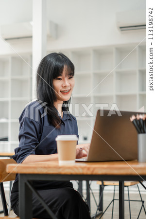 Smiling Asian woman focused on her laptop work in a bright cafe environment with coffee and stationery on the table. 113198742