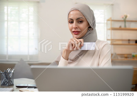 Young happy muslim woman in hijab at home using laptop shopping online with credit card while sitting on desk 113199920