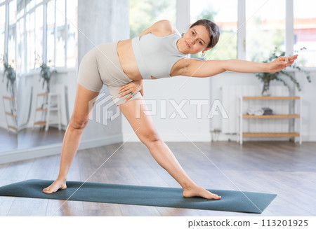 Skilled happy young female teacher demonstrating variation of warrior pose of yoga in training room Skilled happy young female teacher demonstrating variation of warrior pose of yoga in training room 113201925