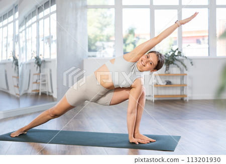 Skilled happy young female teacher demonstrating variation of warrior pose of yoga in training room 113201930