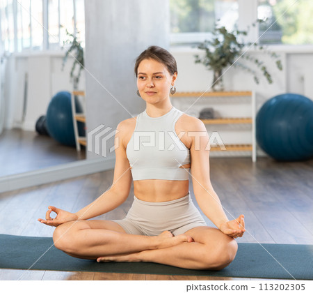 Slender girl doing hatha yoga in lotus position in bright gym 113202305