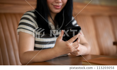 A woman sitting in a coffee shop, using her phone and listening to music on her headphones. 113203058