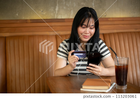 A woman holding a smartphone and a credit card at a table in a coffee shop, paying bills online 113203061