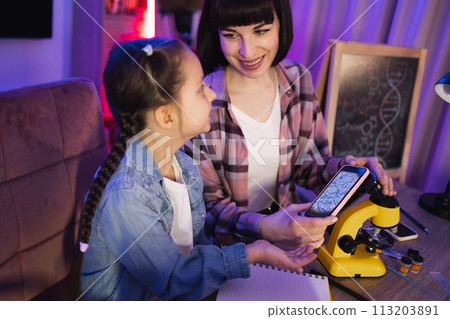Two female sitting at table and doing micrograph with phone at home. Two female sitting at table and doing micrograph with phone at home. 113203891