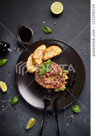 Tuna and avocado tartare with sesame seeds, capers and egg yolk on a dark ceramic plate with baguette bread crouton chips Tuna and avocado tartare with sesame seeds, capers and egg yolk on a dark ceramic plate with baguette bread crouton chips 113206314