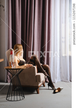 woman relaxes at home sitting in a chair by the window next to a table with whiskey 113207198