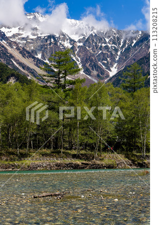 Hikers next to a clear river in front of huge, snowcapped mountains (Hotaka range) 113208215