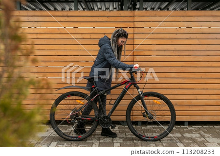 Cute young woman with dreadlocks hairstyle dressed in a winter jacket rides a bicycle Cute young woman with dreadlocks hairstyle dressed in a winter jacket rides a bicycle 113208233