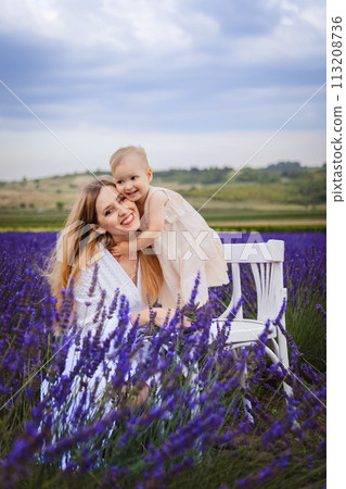 Standing on a white chair in a field of lavender, a happy mother hugs her daughter as she stands on a white chair 113208736