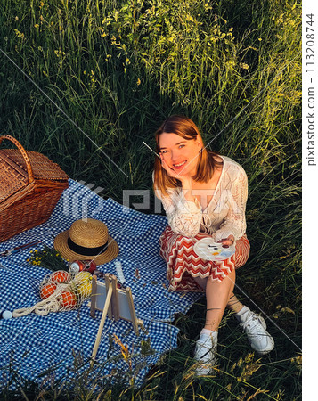 Painting memories: young woman captures sunset on a blue blanket at picnic 113208744