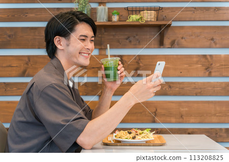 A young man drinking a green smoothie while looking at his smartphone in a cafe (enzymes, healthy, dietary fiber, health food) 113208825