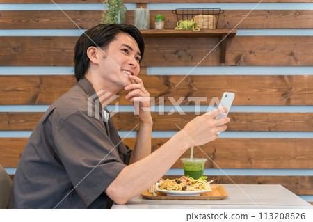 A young man eating/lunch while looking at his smartphone at a cafe 113208826