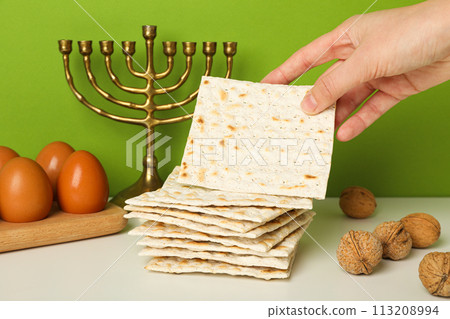 Matzo in hand, nuts, eggs on wooden stand and hanukkiah on green background 113208994