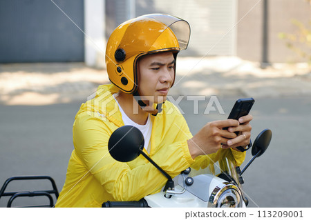 Frowning motorbike taxi driver checking request notifications on smartphone 113209001