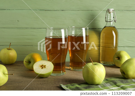 Glasses and bottle with apple cider and green apples on table on wooden green background 113209007