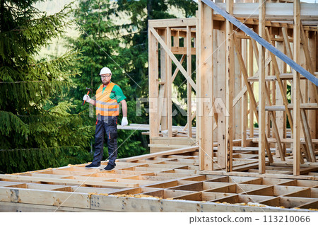 Carpenter constructing wooden frame two-story house near the forest. Bearded man holding hammer, dressed in protective helmet and orange safety vest. Concept of ecological modern construction. Carpenter constructing wooden frame two-story house near the forest. Bearded man holding hammer, dressed in protective helmet and orange safety vest. Concept of ecological modern construction. 113210066