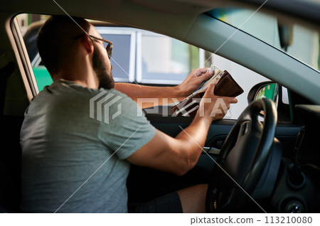 Young man paying for gasoline in cash. Man taking out money from wallet to pay for fuel. High fuel prices concept. 113210080