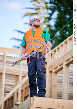 Carpenter building wooden frame two-story house. Bearded man in glasses holding a level, wearing protective helmet, overalls and orange vest. Concept of modern ecological construction. Carpenter building wooden frame two-story house. Bearded man in glasses holding a level, wearing protective helmet, overalls and orange vest. Concept of modern ecological construction. 113210123