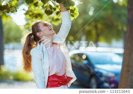smiling trendy woman in pink dress and white jacket in city 113211643