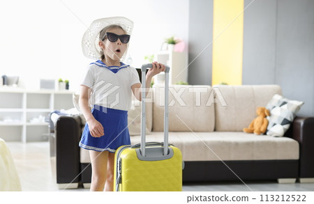 Surprised little girl in sunglasses and hat standing with suitcase in her hands at home Surprised little girl in sunglasses and hat standing with suitcase in her hands at home 113212522